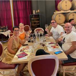 Group enjoying wine tasting at a table with wine bottles and barrels in the background.