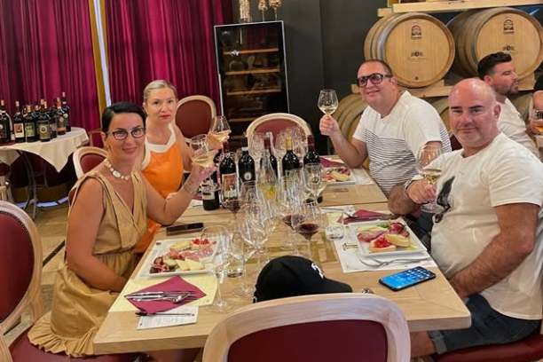 Group enjoying wine tasting at a table with wine bottles and barrels in the background.