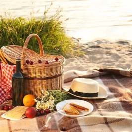 Picnic setup with basket, wine, fruits, and hat on a beach blanket by the water.