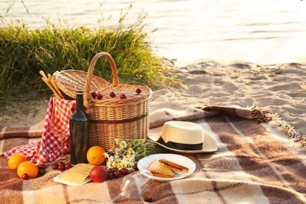 Picnic setup with basket, wine, fruits, and hat on a beach blanket by the water.