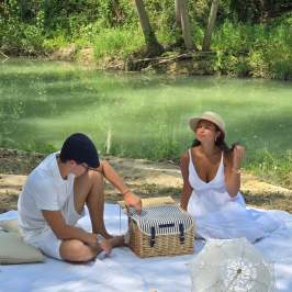 Two people in white outfits have a picnic by a river with a basket and umbrella.