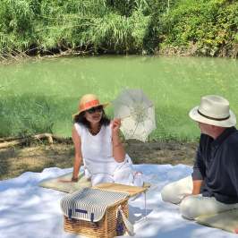 Two people having a picnic by a green pond with hats and a parasol.